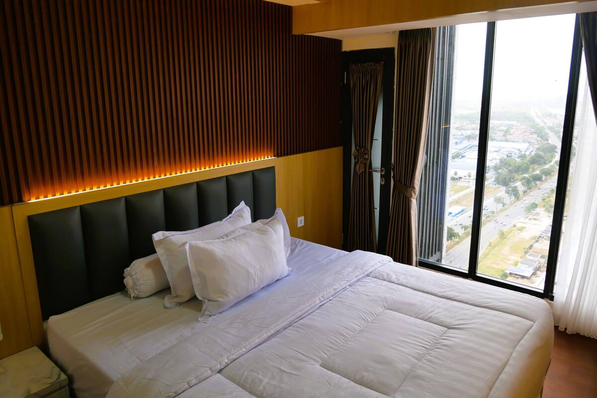 A hotel room with a large bed, a headboard with vertical slats, and a view of a cityscape through the window.