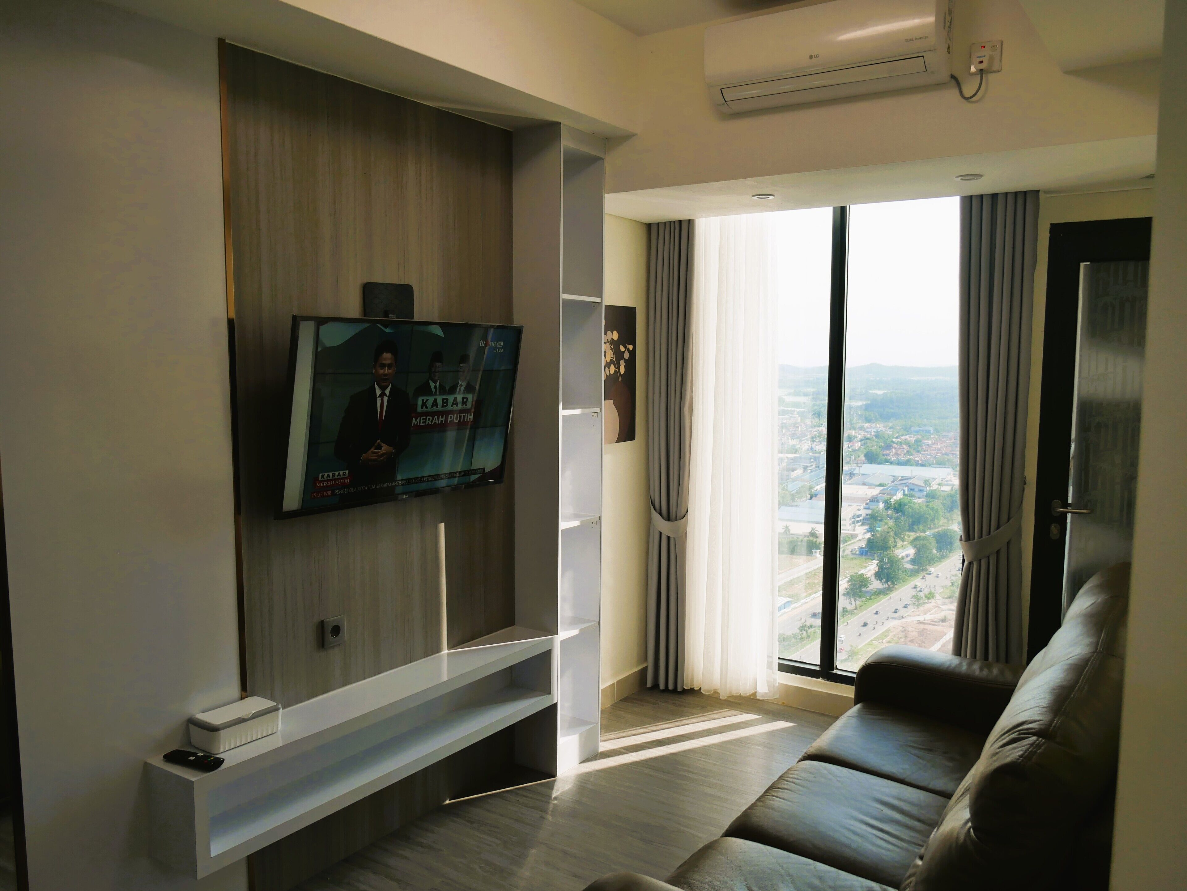 A modern living room with a flat-screen TV mounted on the wall, a leather sofa, and a large window offering a city view.