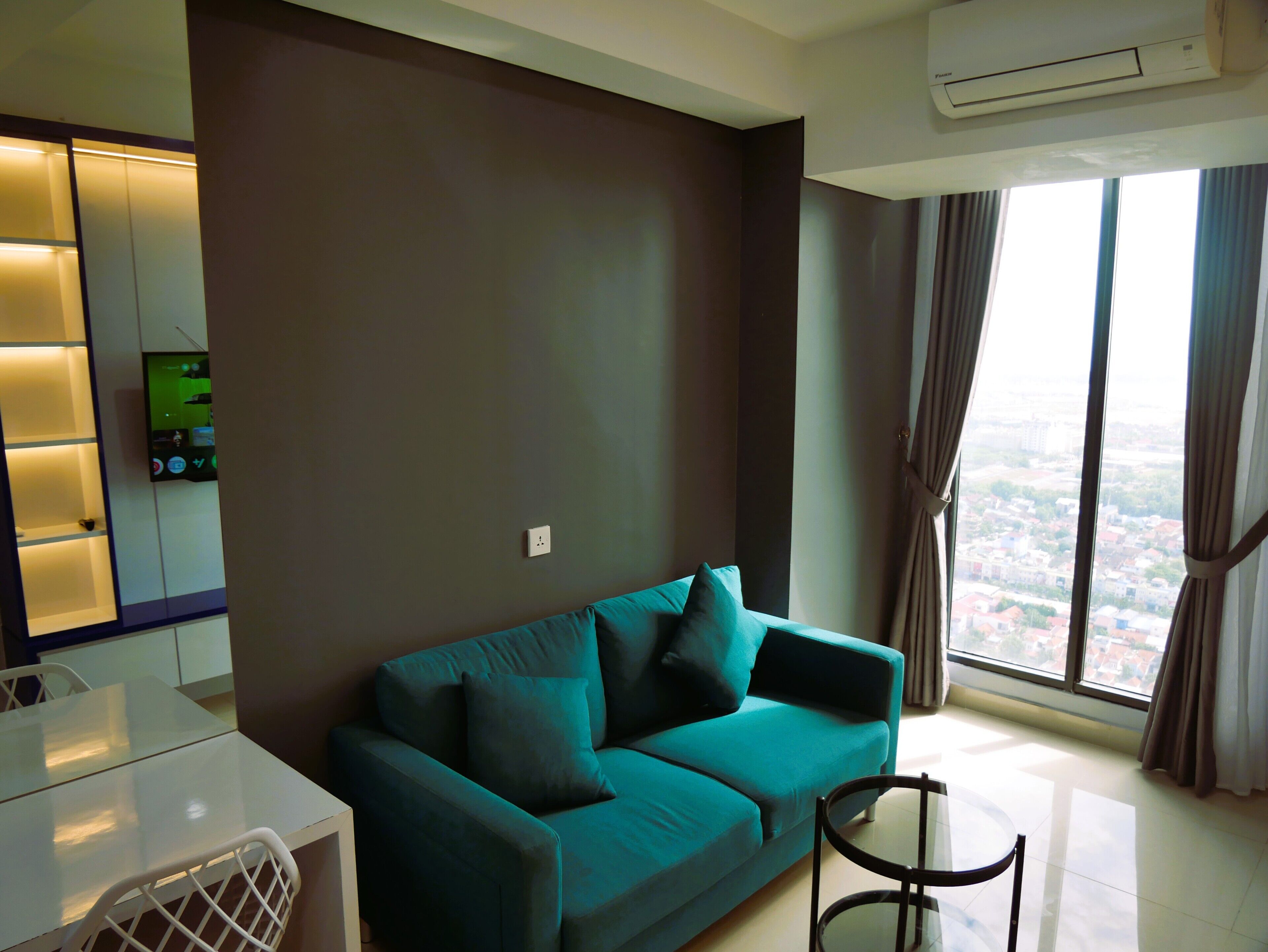 A modern living room with a teal sofa, a glass table, and a city view through large windows.