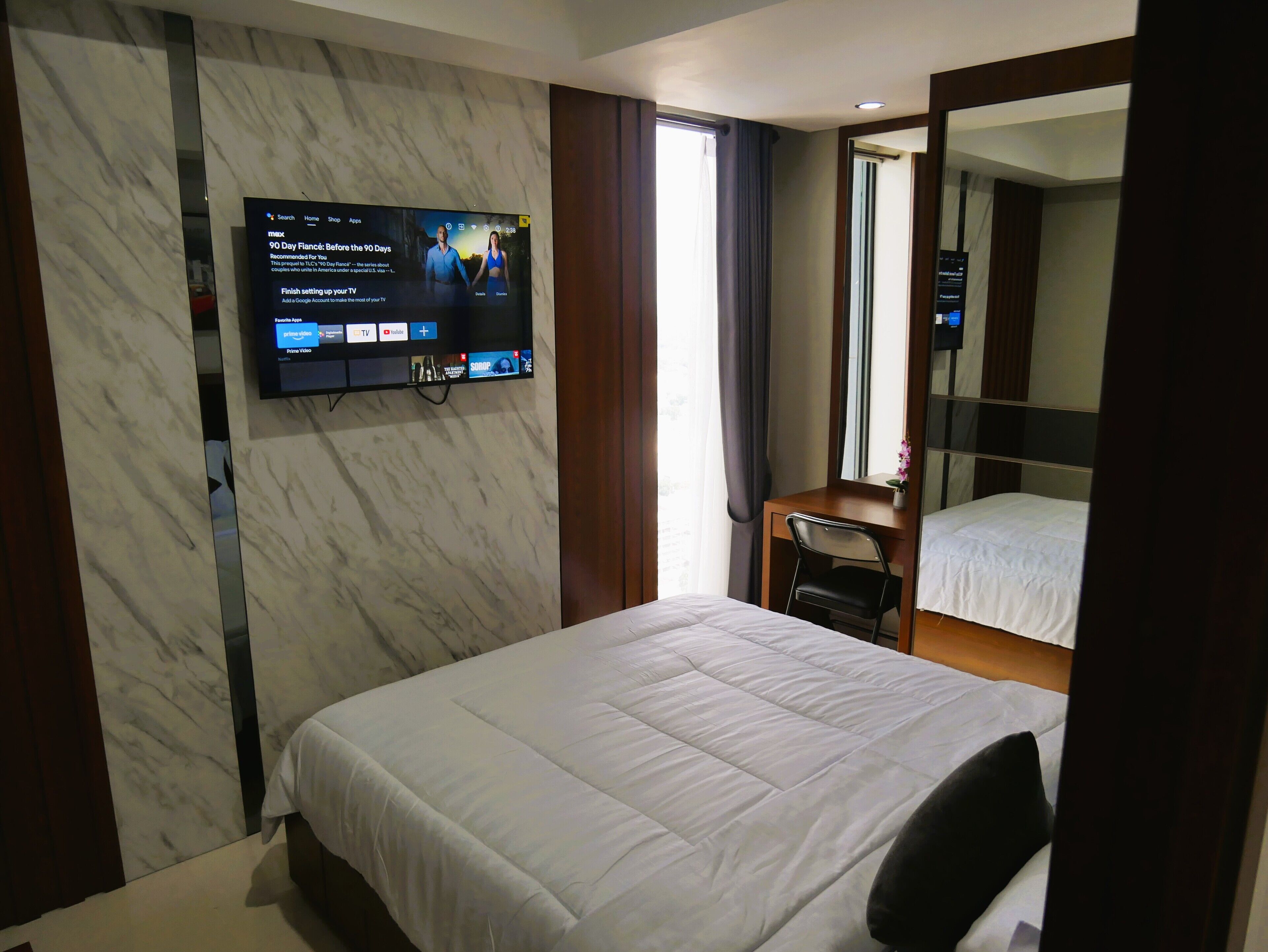 A hotel room with a large flat-screen TV mounted on the wall, a bed with white bedding, a small desk with a chair, and a large mirror above the bed.