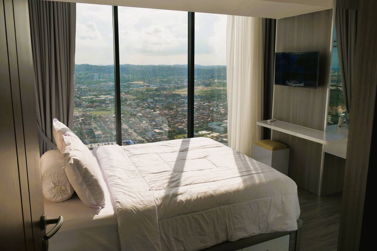 Une chambre dotée d’un grand lit, d’un téléviseur fixé au mur et offrant une vue sur le paysage urbain grâce à de grandes fenêtres.