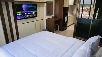 Habitación de hotel con cama, televisión fija en la pared mostrando varios canales, escritorio con silla y vista a la ciudad a través de un ventanal grande.