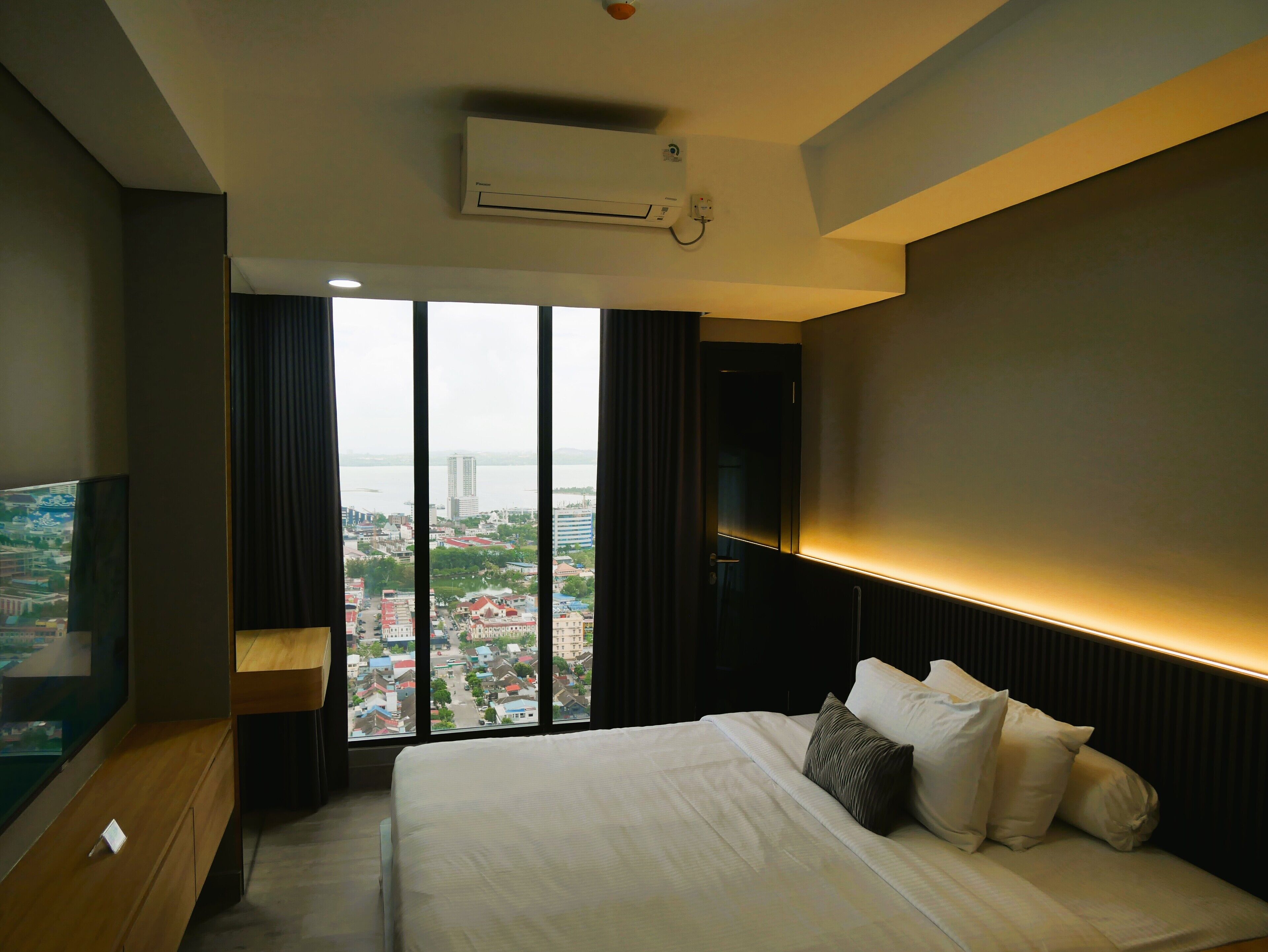 Habitación de hotel con una cama grande, televisión, vistas de un paisaje urbano y un equipo de aire acondicionado.
