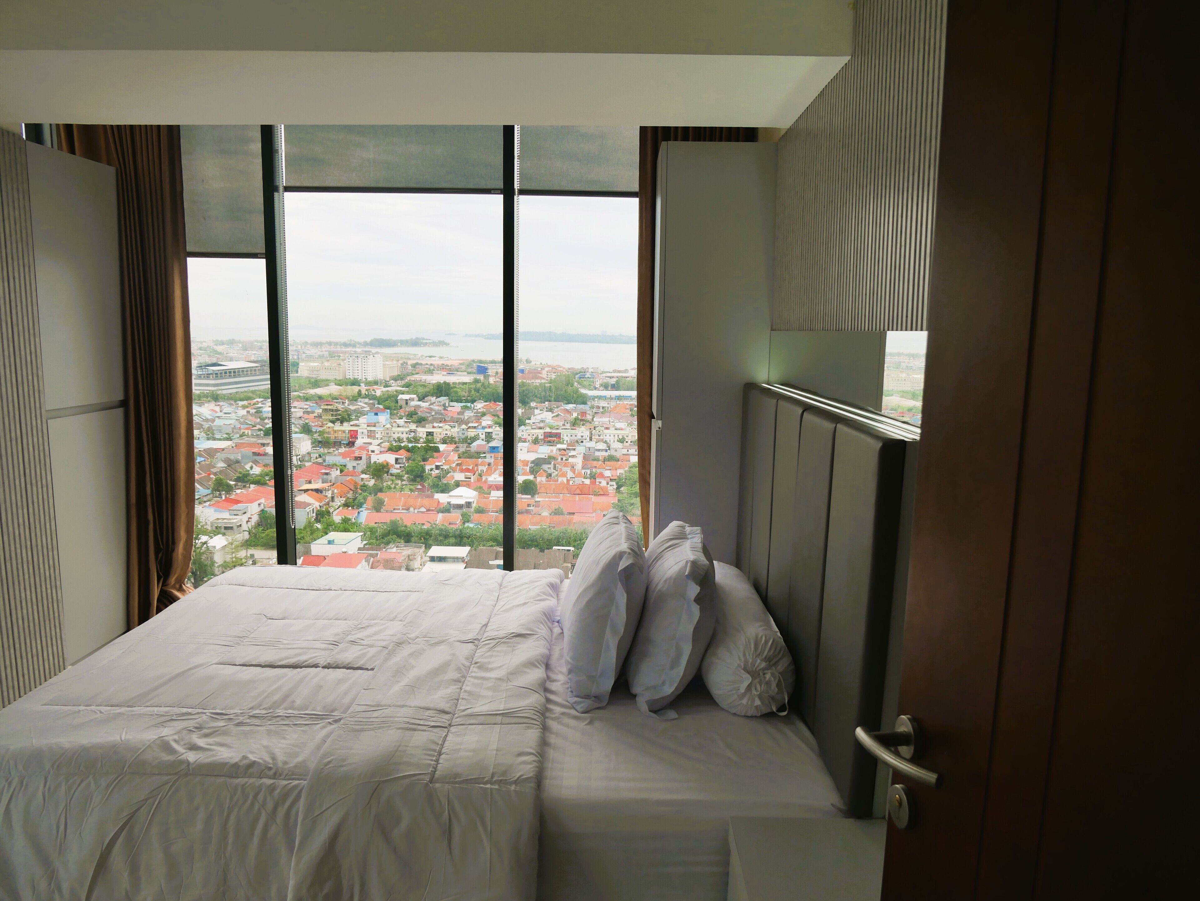 Une chambre d’hôtel avec un grand lit, une vue sur le paysage urbain grâce à de grandes fenêtres du sol au plafond et une tête de lit en bois.
