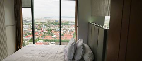 Une chambre d’hôtel avec un grand lit, une vue sur le paysage urbain grâce à de grandes fenêtres du sol au plafond et une tête de lit en bois.