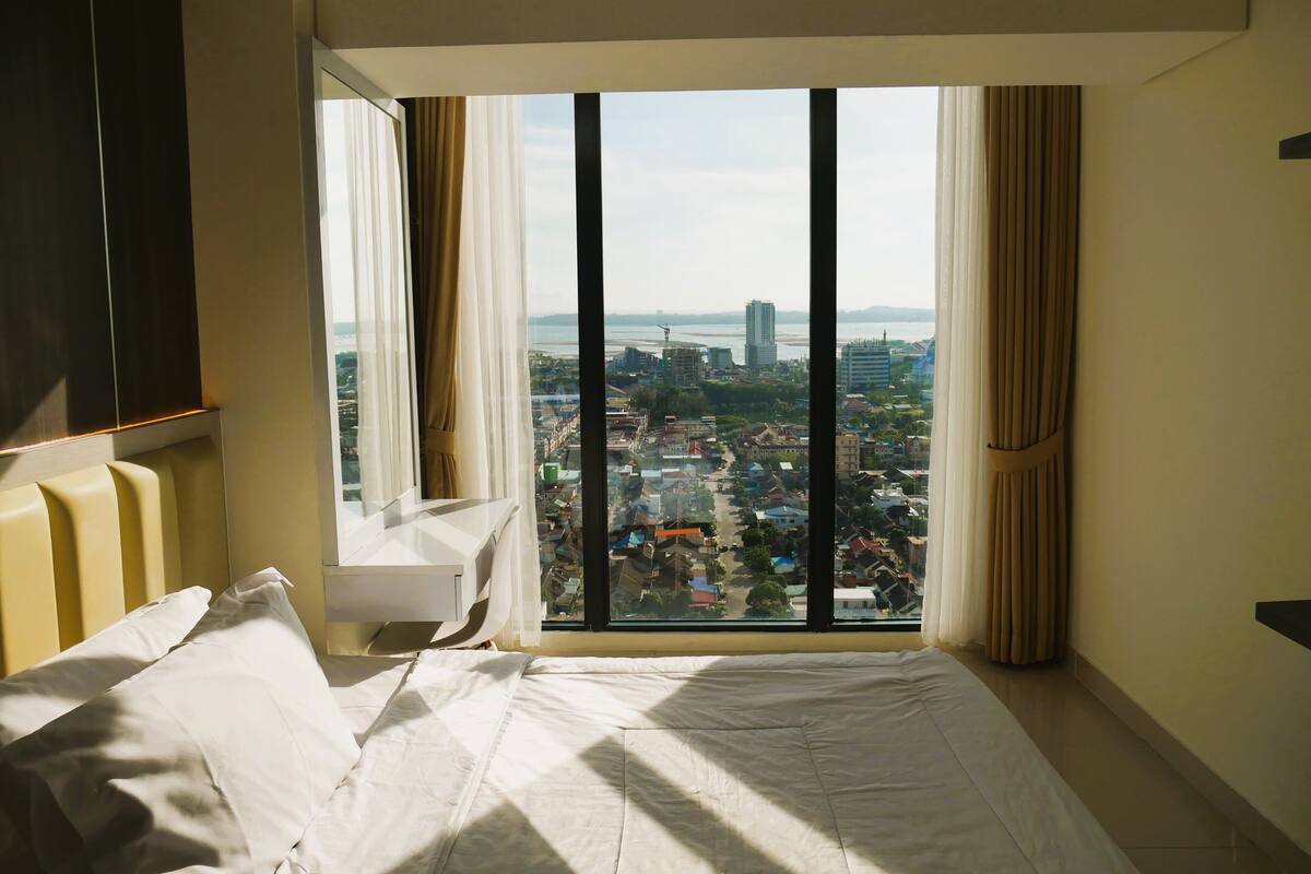 A bedroom with a large window, a bed, a bedside table, and a wardrobe.