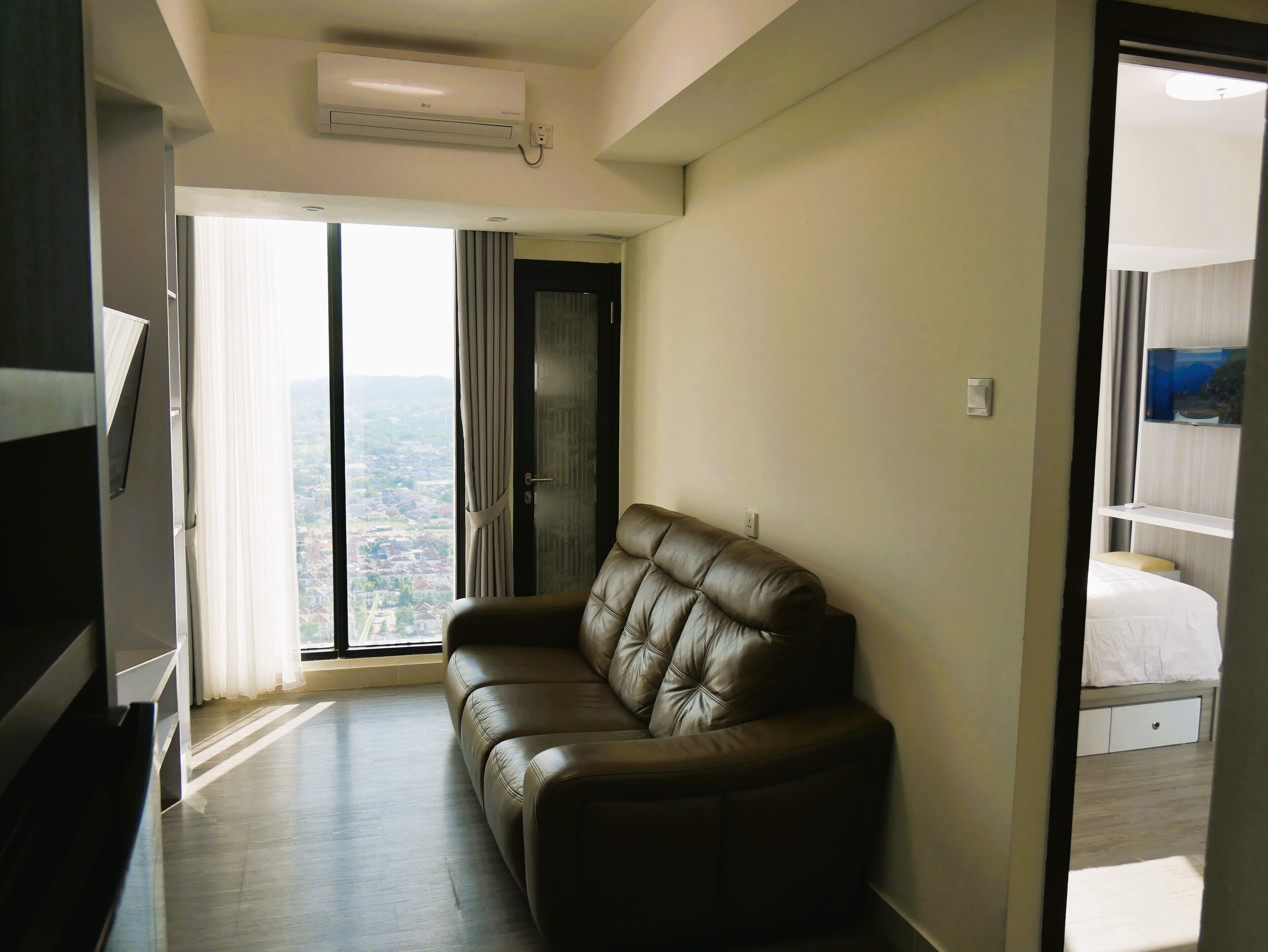 A modern living room with a leather sofa, a flat-screen TV, and a large window offering a city view.
