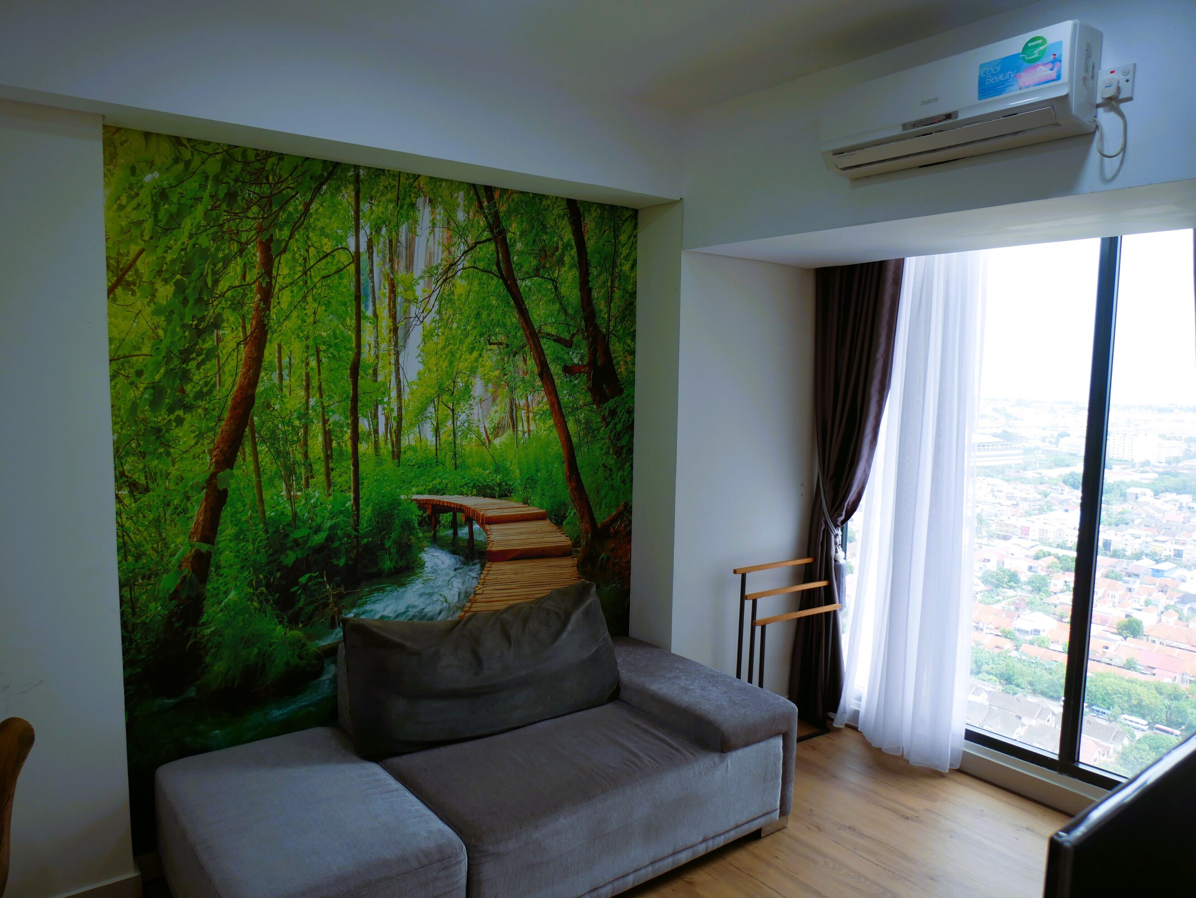 A living room with a grey sofa, a large forest mural on the wall, a wooden bench, and a view of a city through a window.