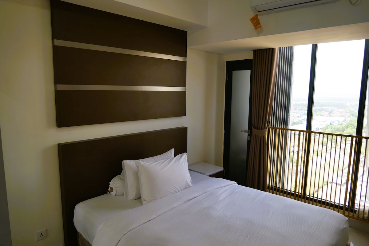 A hotel room with a large bed, a wall-mounted air conditioner, and a window with a view of the city.
