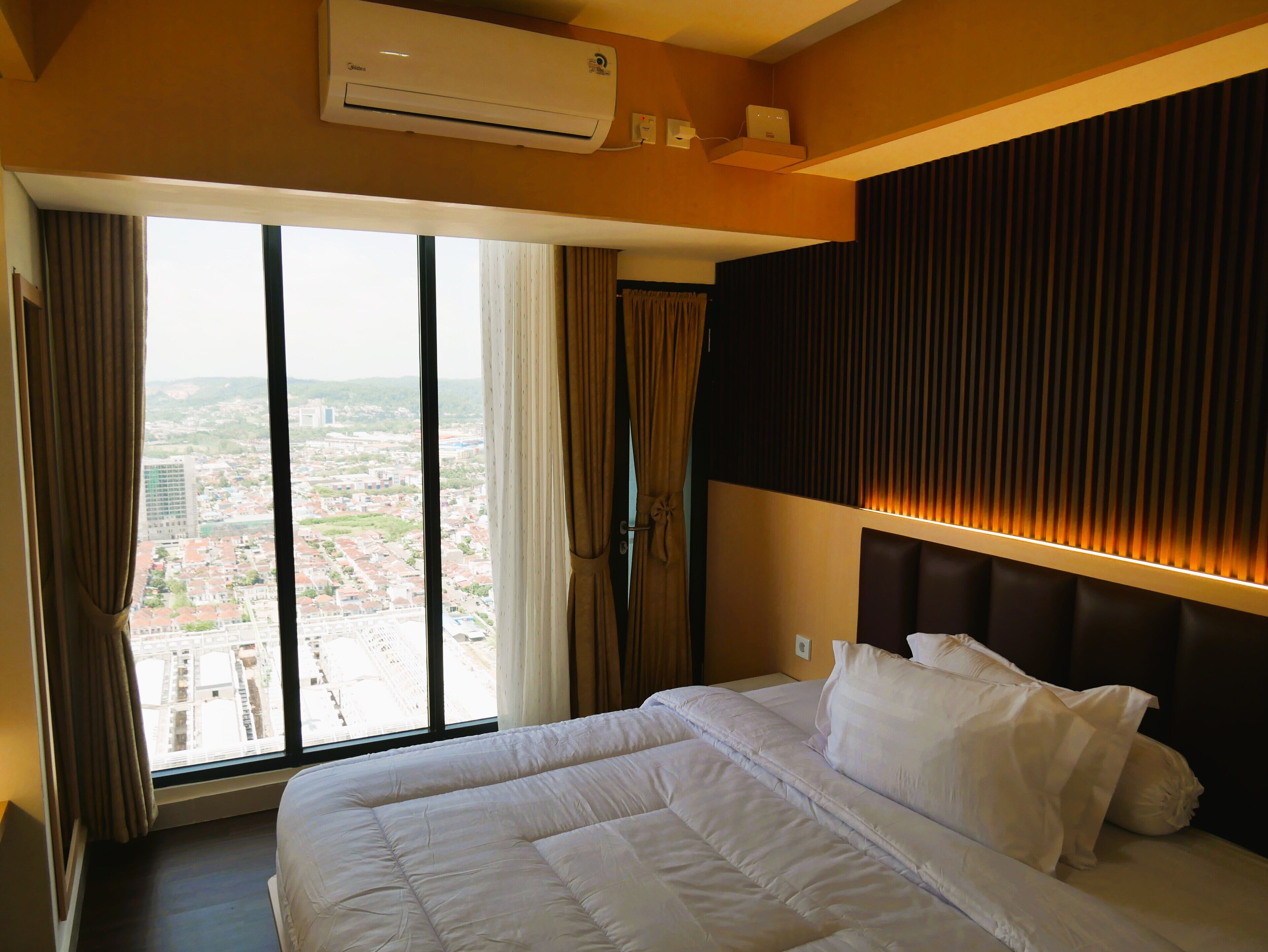A hotel room with a large bed, an air conditioning unit, a view of a cityscape through a large window, and a modern headboard with built-in lighting.