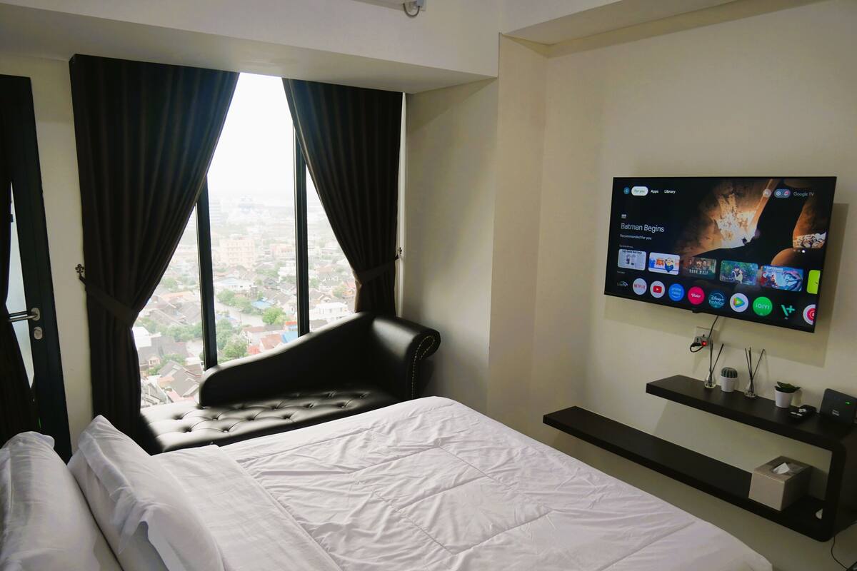 Ett hotellrum med en säng, ett fönster med gardiner, en platt-TV som är monterad på väggen och visar gränssnittet för en streamingtjänst, och en liten hylla med dekorativa föremål.