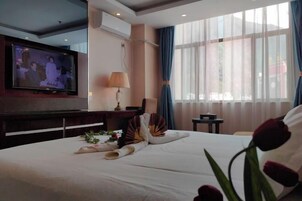 A hotel room with a bed, a television mounted on the wall, a small table with a lamp, and a window with curtains.