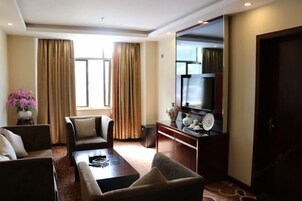 Habitación de hotel con sofá, sillón y una mesita de centro. Hay un televisor sobre un mueble, un florero con flores y una ventana con cortinas.
