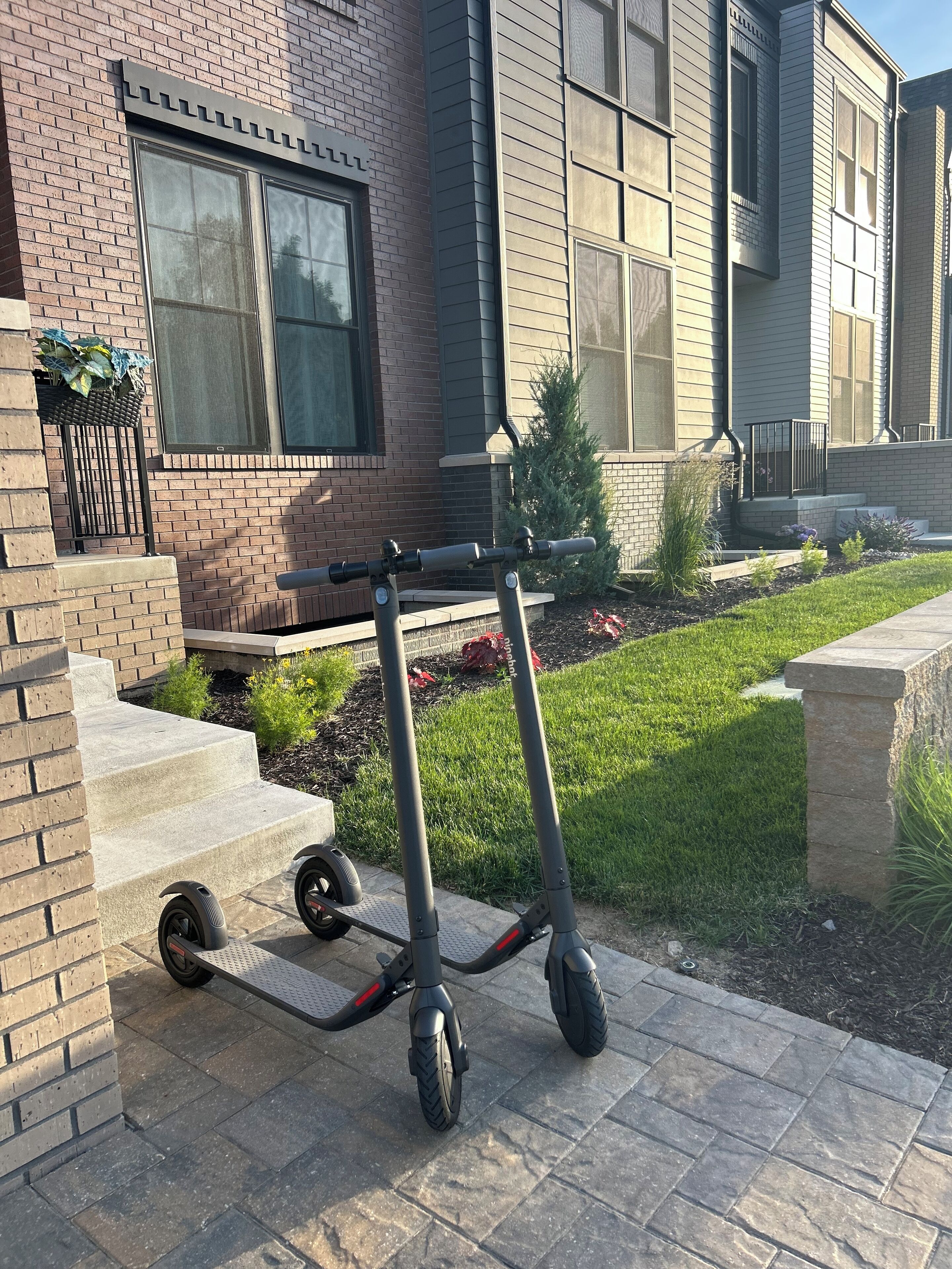 Un scooter eléctrico de dos ruedas estacionado en un área pavimentada frente a un edificio con fachada de ladrillo y revestimiento, escalones y un pequeño jardín con plantas.
