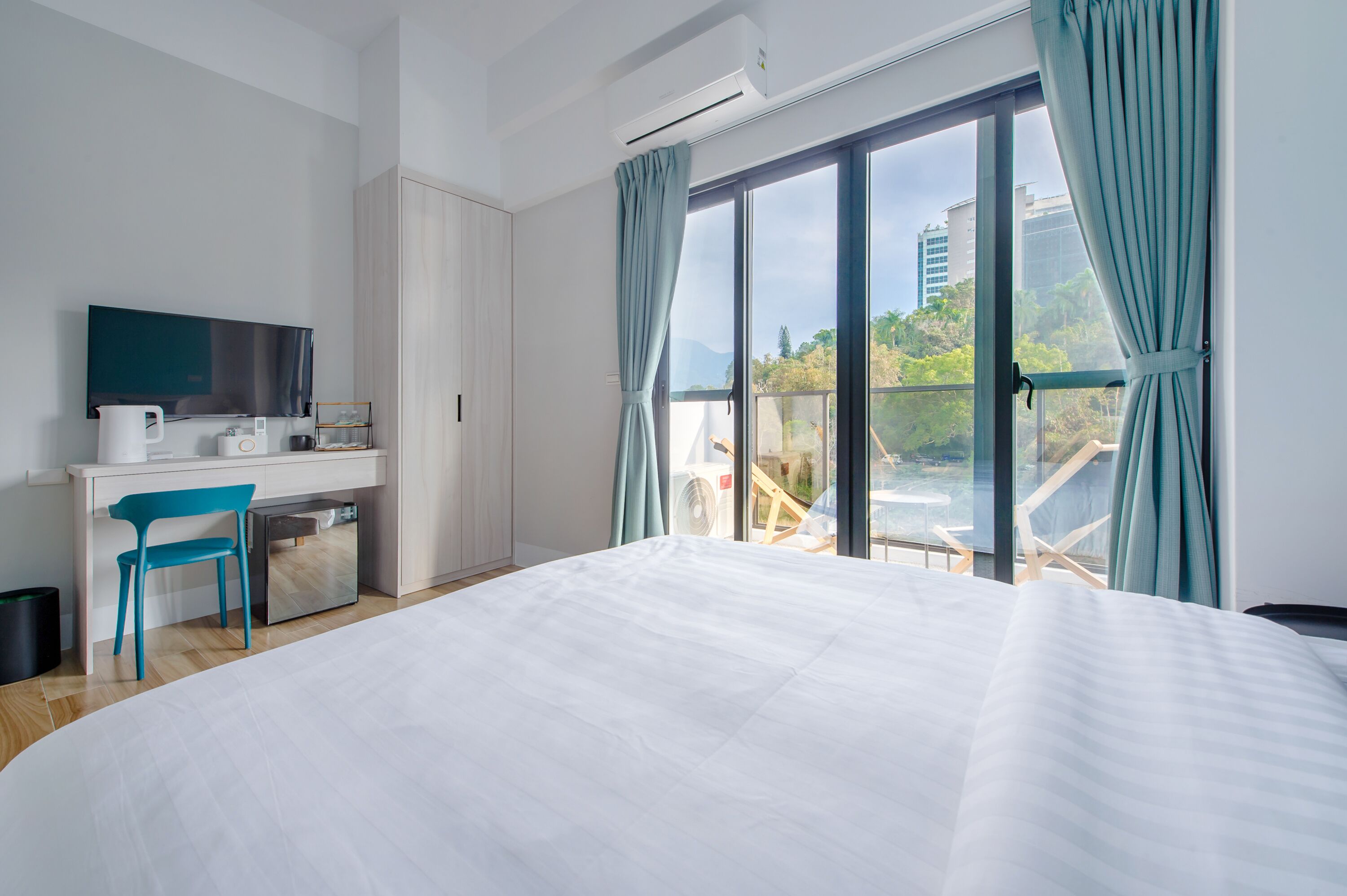 Una habitación de hotel moderna con una cama grande, un escritorio con un televisor de pantalla plana, una silla y un balcón con vistas a la ciudad.