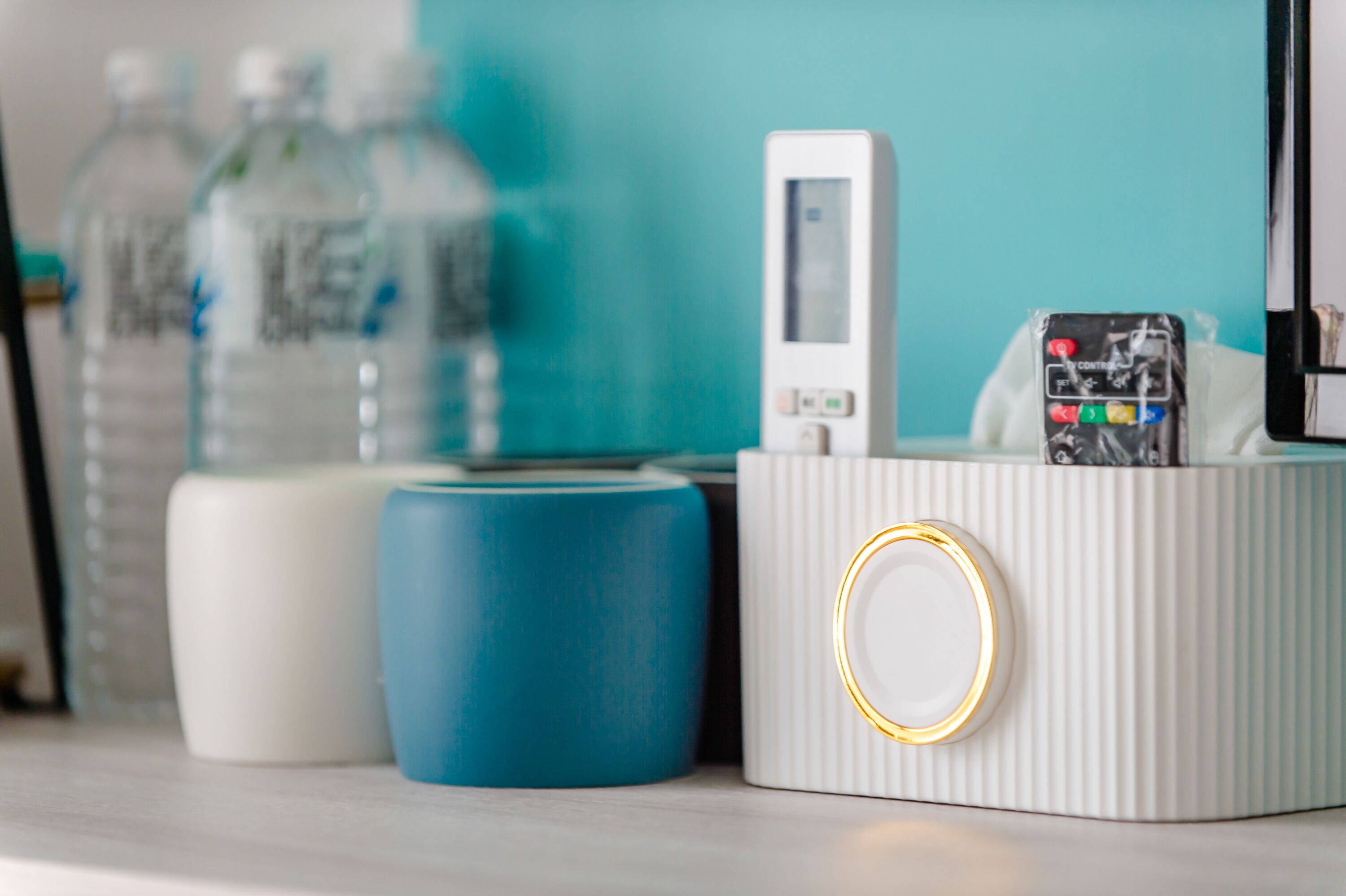 Uma mesa de cabeceira branca com uma luminária branca, um objeto cilíndrico azul e um recipiente branco com um aro dourado.