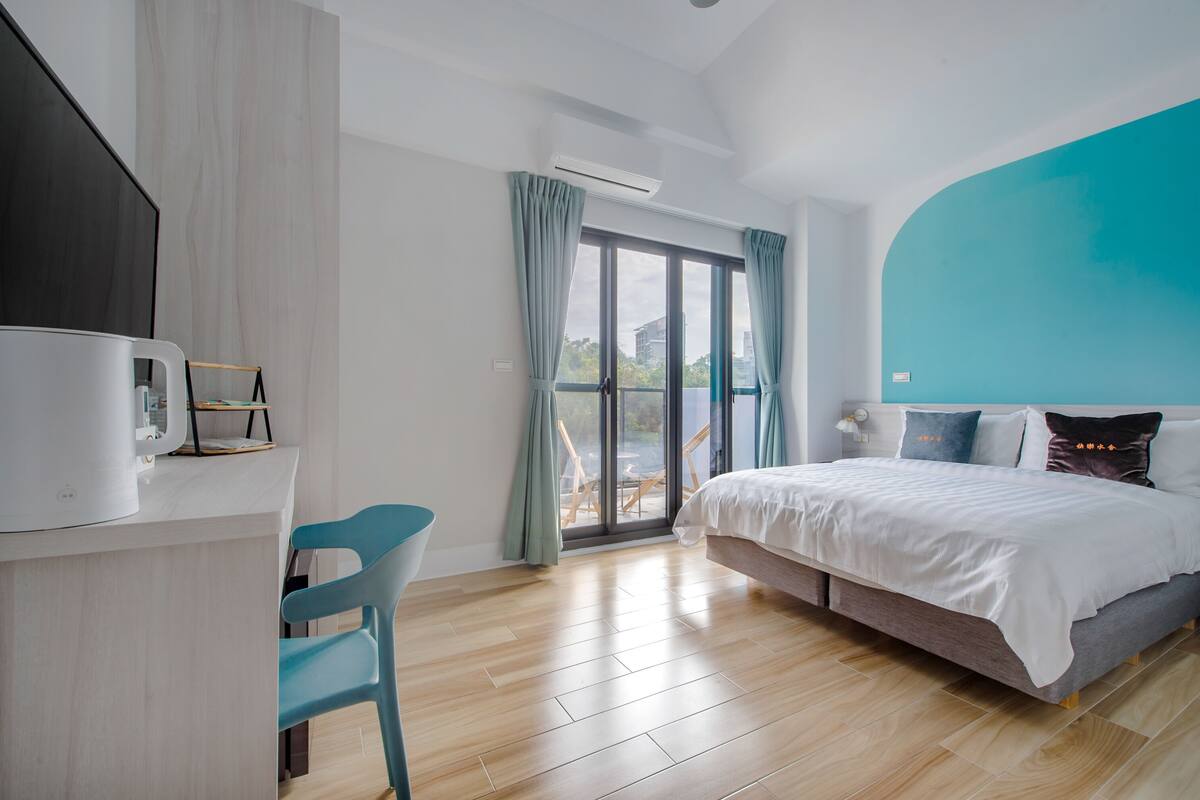 A modern hotel room with a large bed, a desk with a chair, and a balcony with a view of the city.