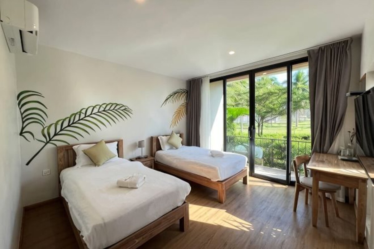 A hotel room with two beds, a desk, and a large window with a view of greenery.