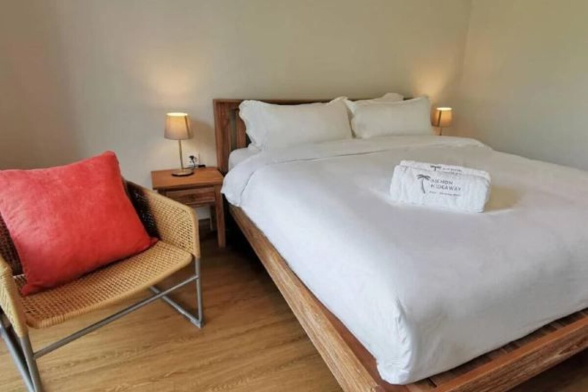 A hotel room with a single bed, a nightstand with a lamp, a chair with a red cushion, and a wooden floor.