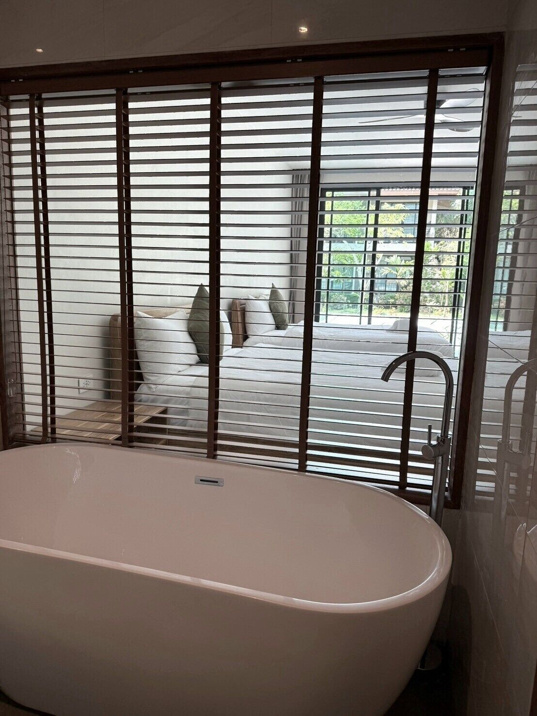 Un baño moderno con una bañera independiente, una ventana con persianas horizontales y vistas a un patio exterior.