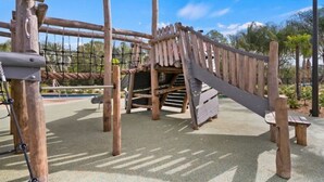 A wooden play structure with a slide and climbing elements, surrounded by a paved area and palm trees. - Storey Lake Water Park Resort Condo next to Disney (Kissimmee)