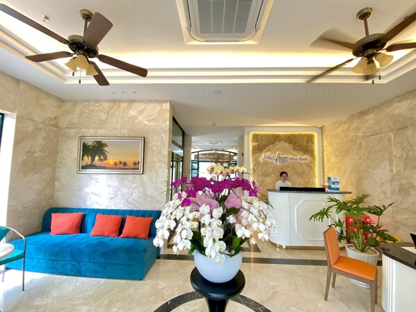 A hotel lobby with a reception desk, a blue sofa, a white vase with orchids, ceiling fans, and a framed picture on the wall.