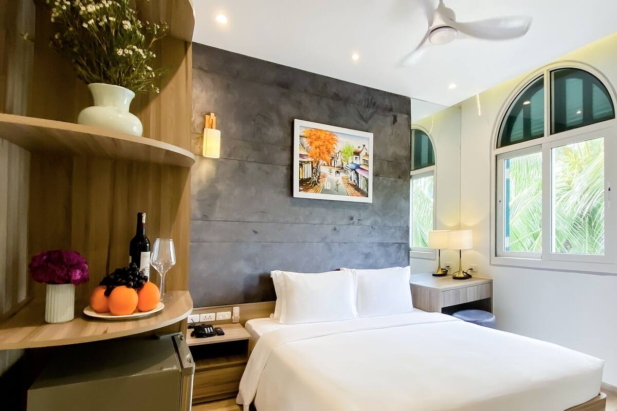 Una habitación de hotel moderna con cama, escritorio, refrigerador, ventilador de techo, un cuadro y ventana con vista.