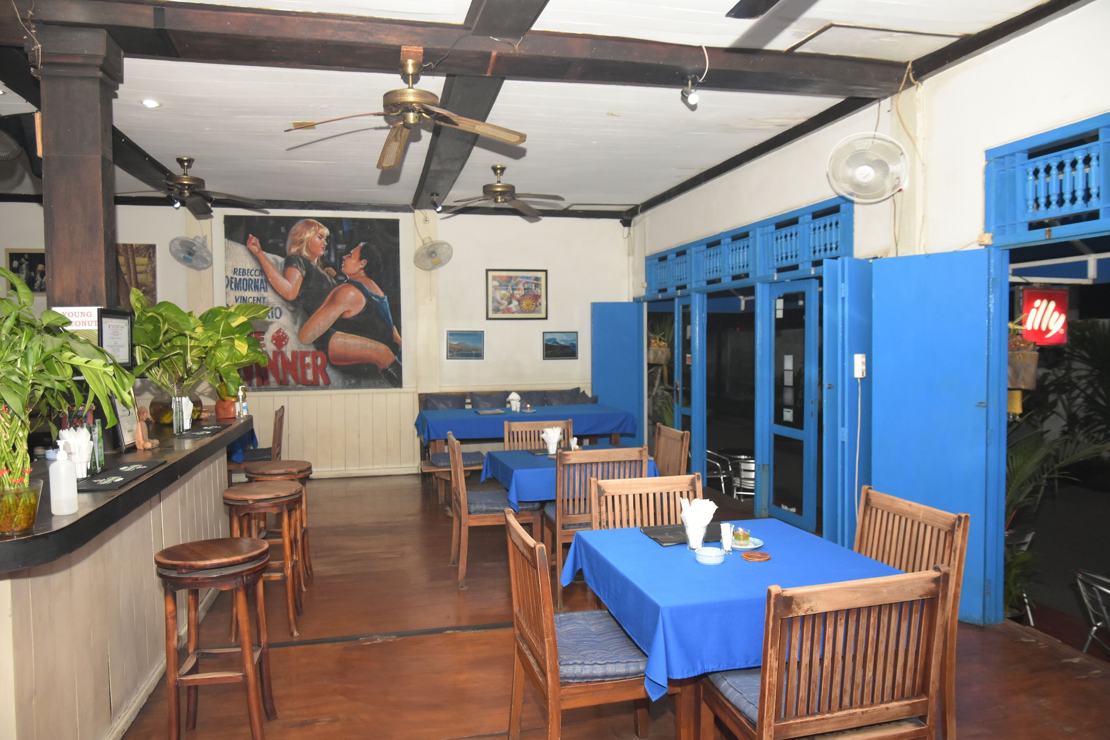 A restaurant interior with wooden tables and chairs, blue tablecloths, ceiling fans, and a large poster on the wall.