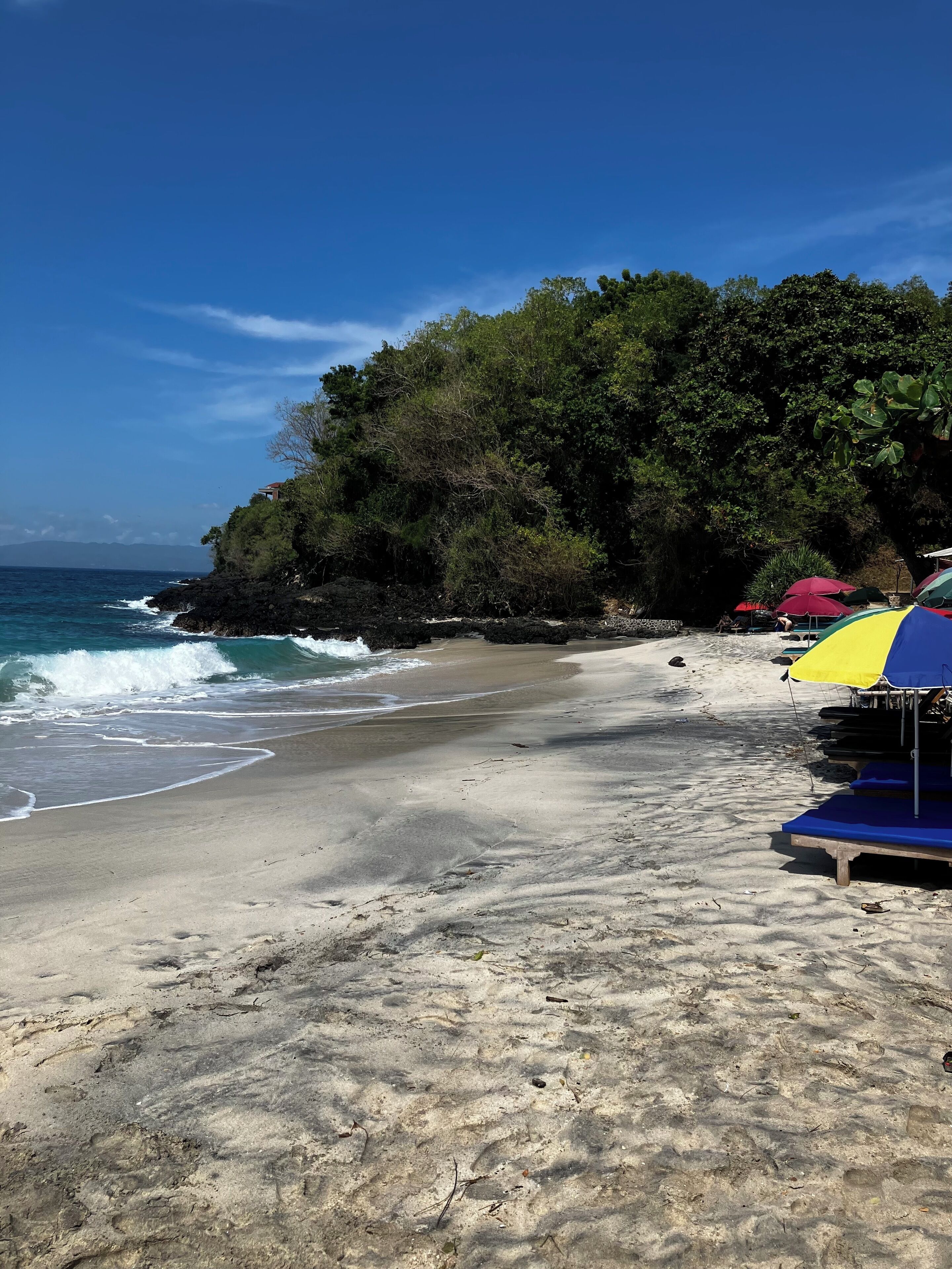 Ein Strand mit felsigem Ufer, blauem Himmel und einigen Sonnenschirmen.