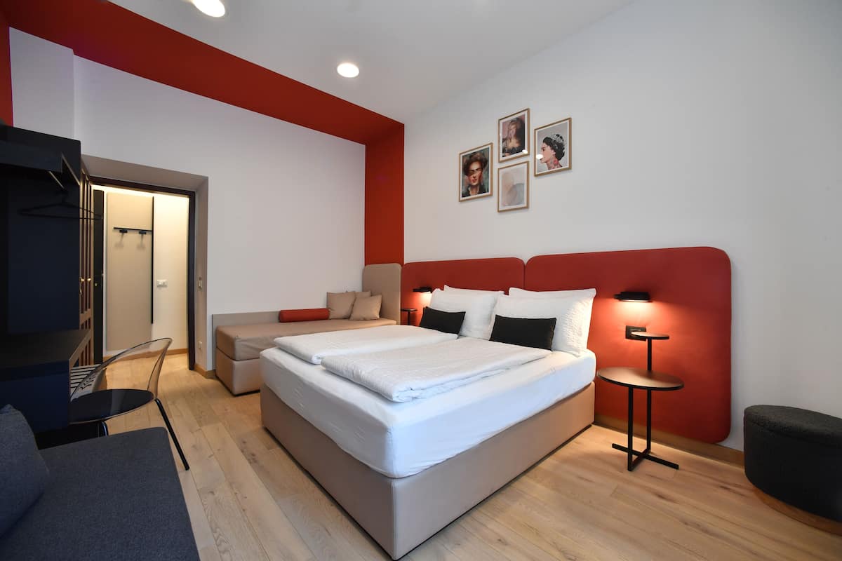 A modern bedroom with a large bed, a desk, and a chair. There is a small table with a lamp and a picture on the wall.