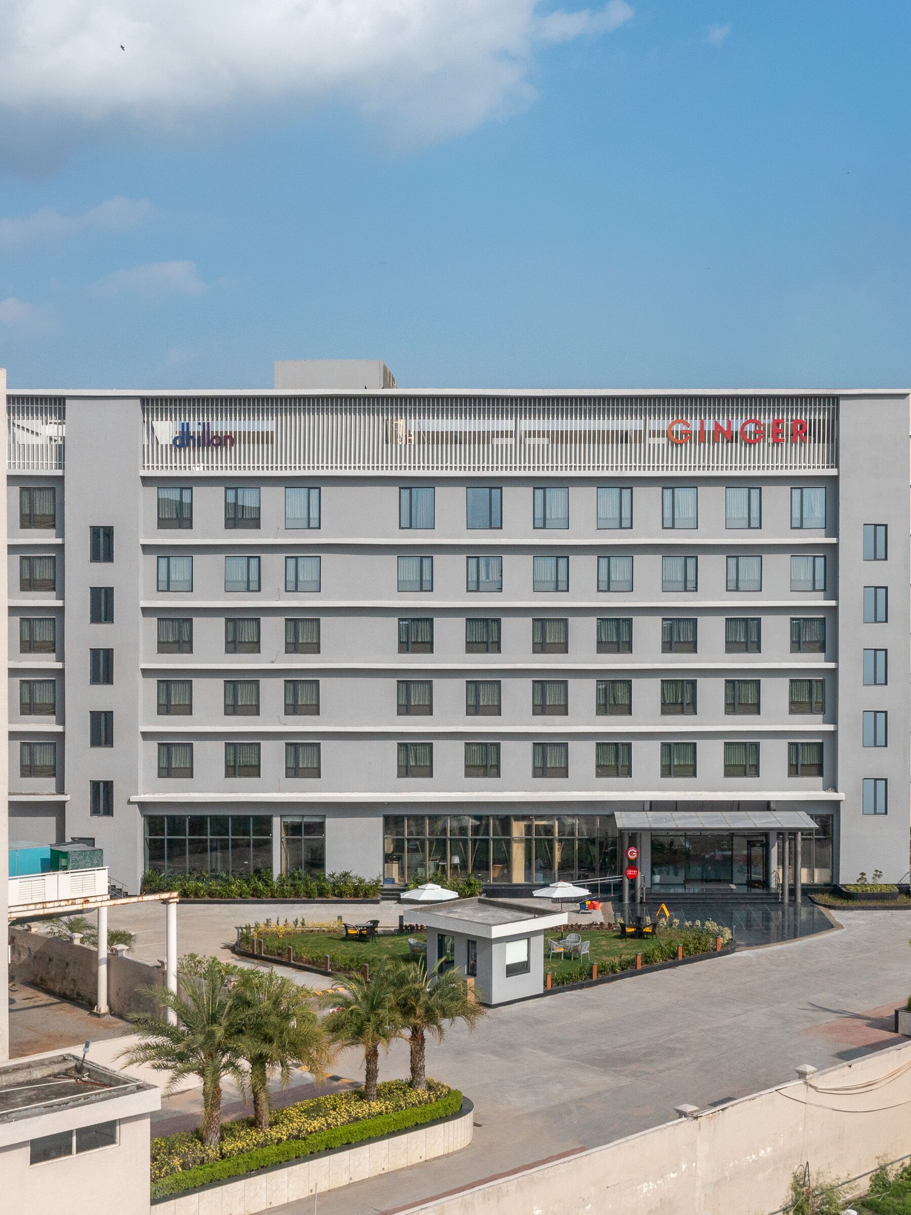 Un hotel moderno con fachada plana, varios balcones y un área de entrada cubierta con mobiliario exterior.