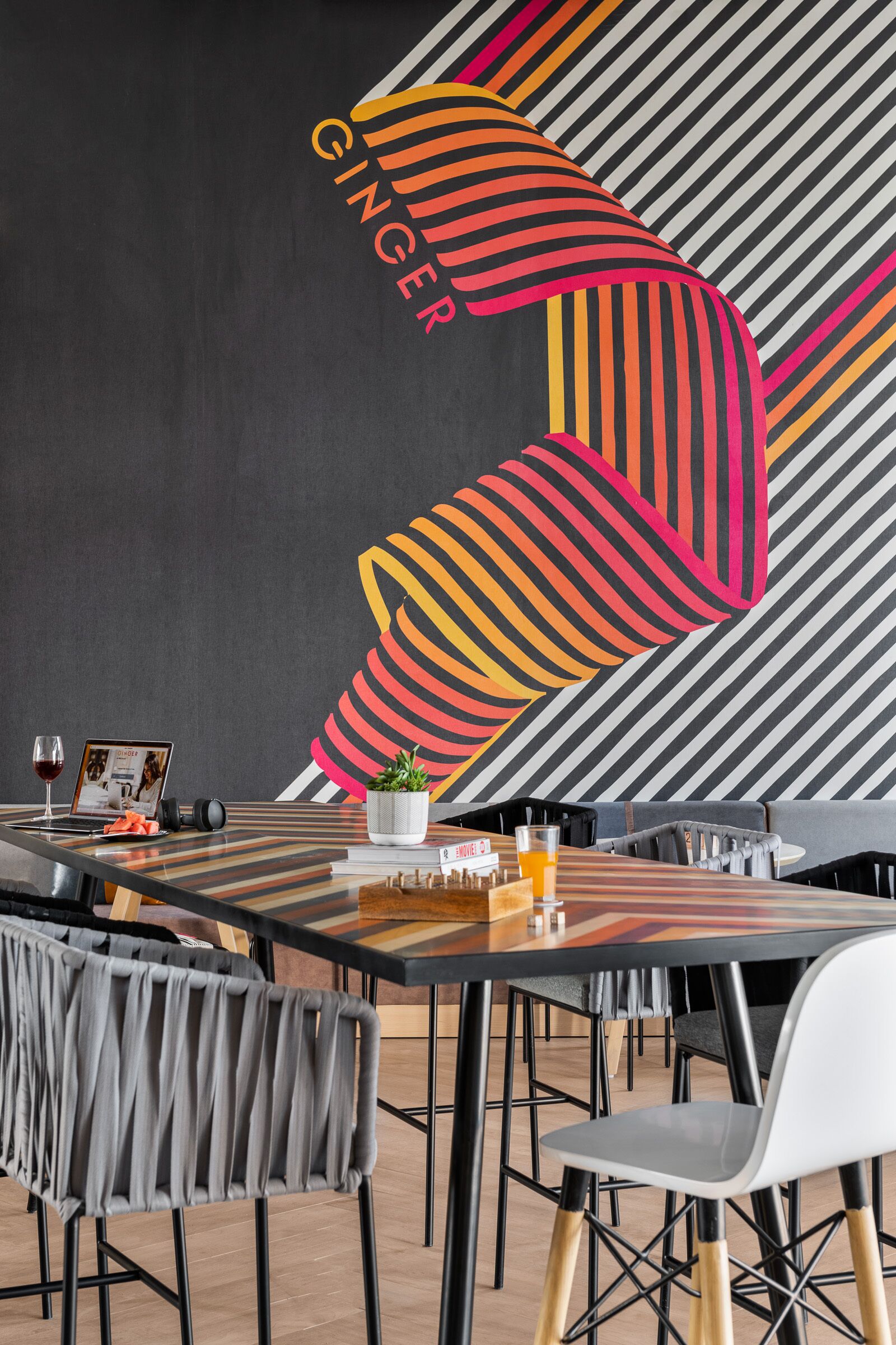 Un espace repas moderne avec des motifs rayés sur les murs, un ordinateur portable, un verre à vin et une plante en pot sur la table.