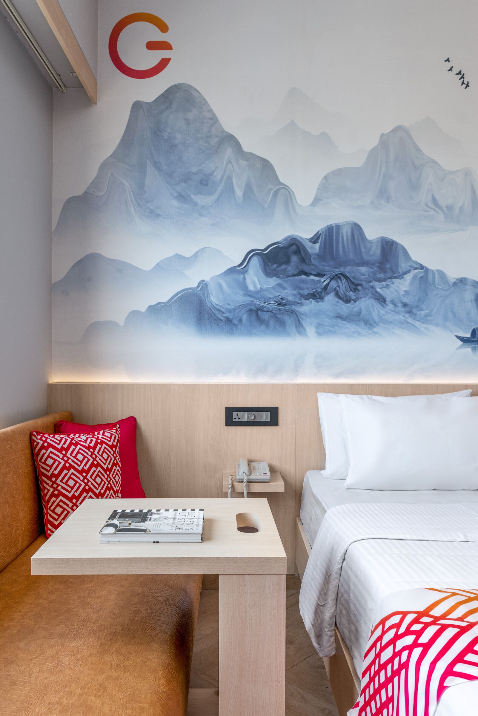 Habitación de hotel con cama, una mesita con una revista, un cojín estampado rojo y una pared con un cuadro de un paisaje montañoso.