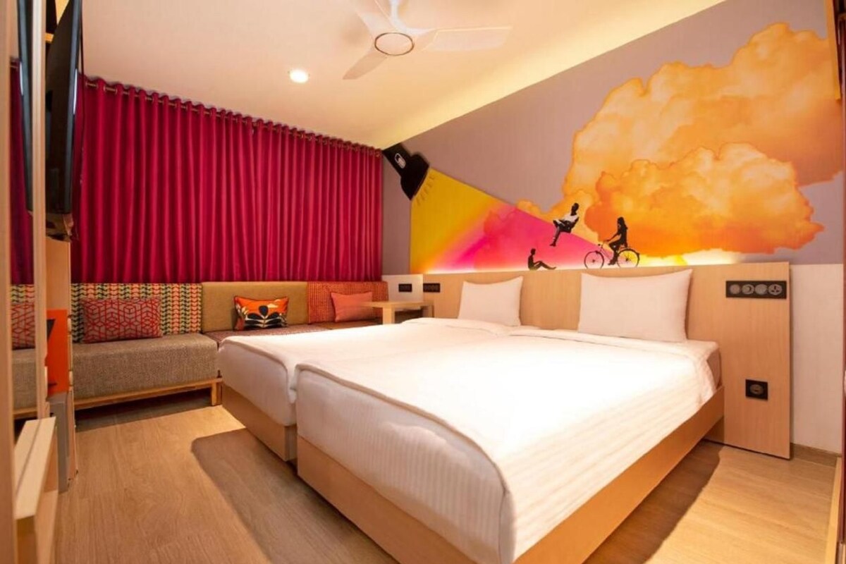 A modern hotel room with a large bed, a sofa, a ceiling fan, and a mural of a cyclist on a rainbow.