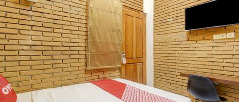 Habitación de hotel con pared de ladrillo, cama con colcha roja y blanca, puerta de madera, televisor montado en la pared y una silla.