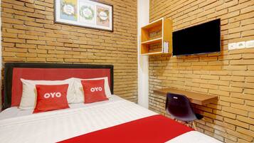 A hotel room with a brick wall, a bed with red and white bedding, a mounted TV, and a small wooden shelf.
