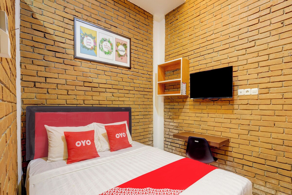 A hotel room with a brick wall, a bed with red and white bedding, a TV mounted on the wall, and a shelf with a few items.
