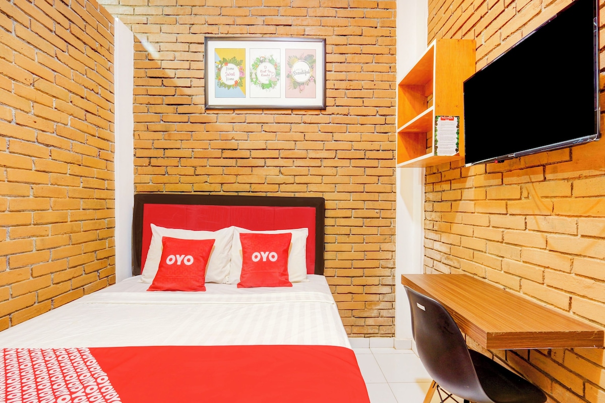 A hotel room with a brick wall, a bed with red and white bedding, a TV mounted on the wall, and a wooden desk.