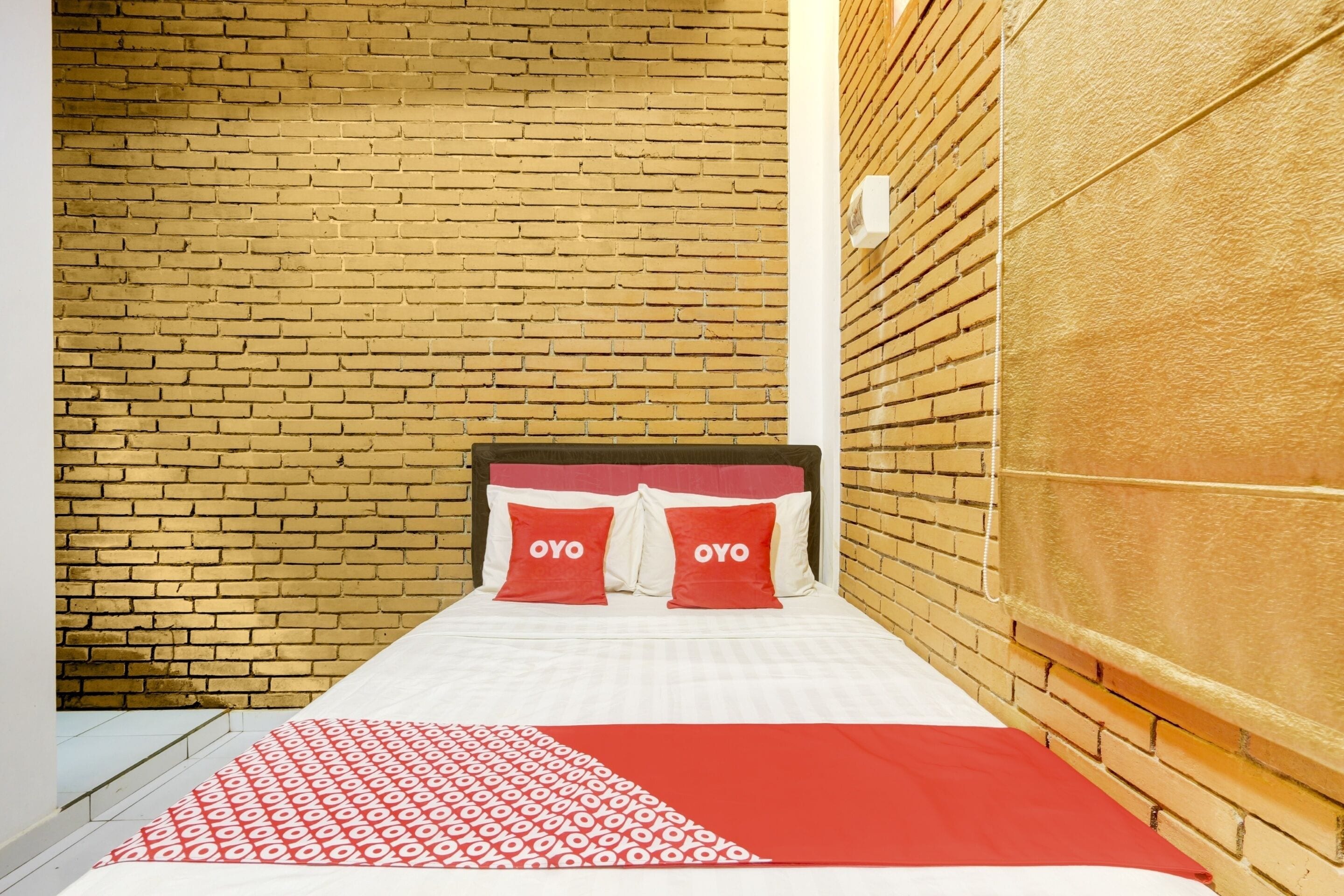 Una cama con una colcha blanca y roja estampada, dos almohadas rojas con la palabra "OYO" y una pared de ladrillo detrás de la cama.