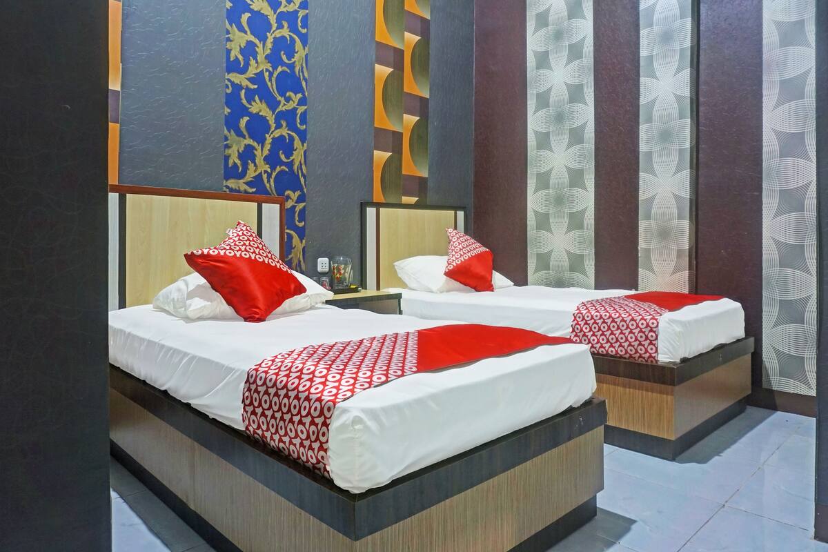 A hotel room with two beds, each with white bedding and red accents. The room features a modern design with patterned wallpaper and a wooden headboard.
