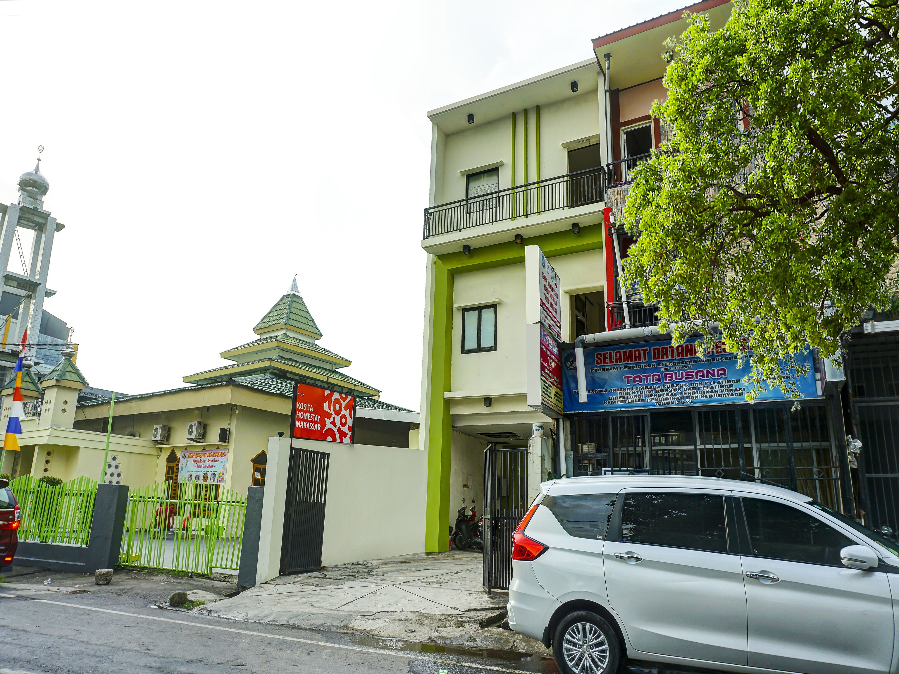 A multi-story building with a green and white facade, a signboard advertising a homestay program, and a white car parked in front.