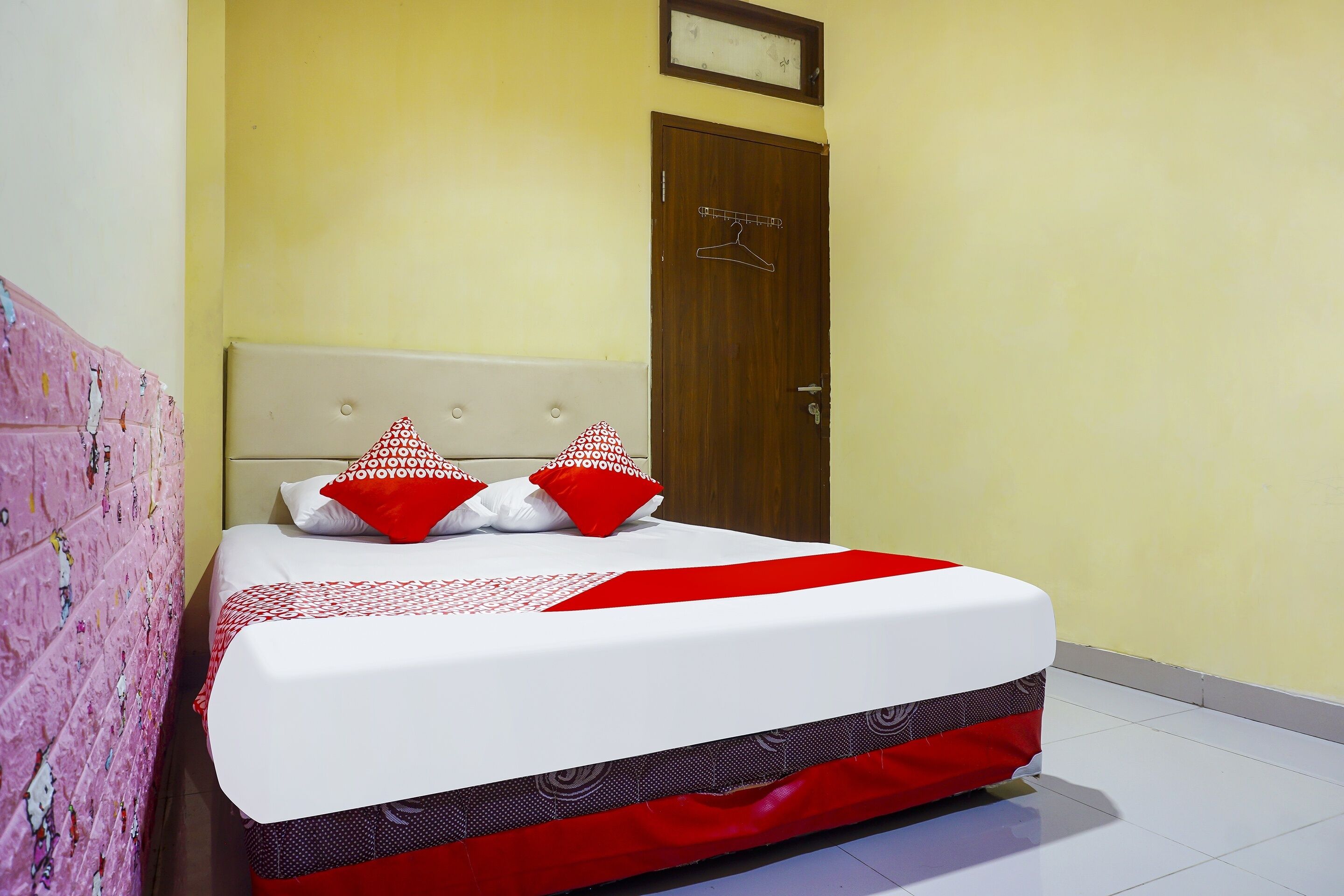 Una cama bien hecha con una colcha blanca y roja, dos almohadas rojas y blancas, y una puerta de madera con un perchero.