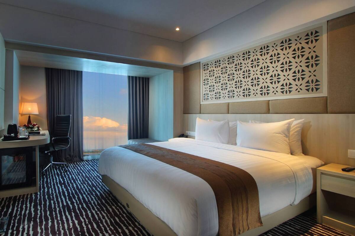 A hotel room with a large bed, a desk with a lamp, a TV, and a view of the sky.