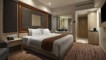 Habitación de hotel con una cama grande, una mesita de noche con teléfono y papeles, una silla, un televisor montado en la pared y un baño visible a través de una puerta abierta.