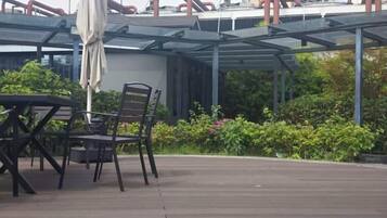 An outdoor patio with a table and chairs, a covered seating area, and a view of greenery and buildings in the background.