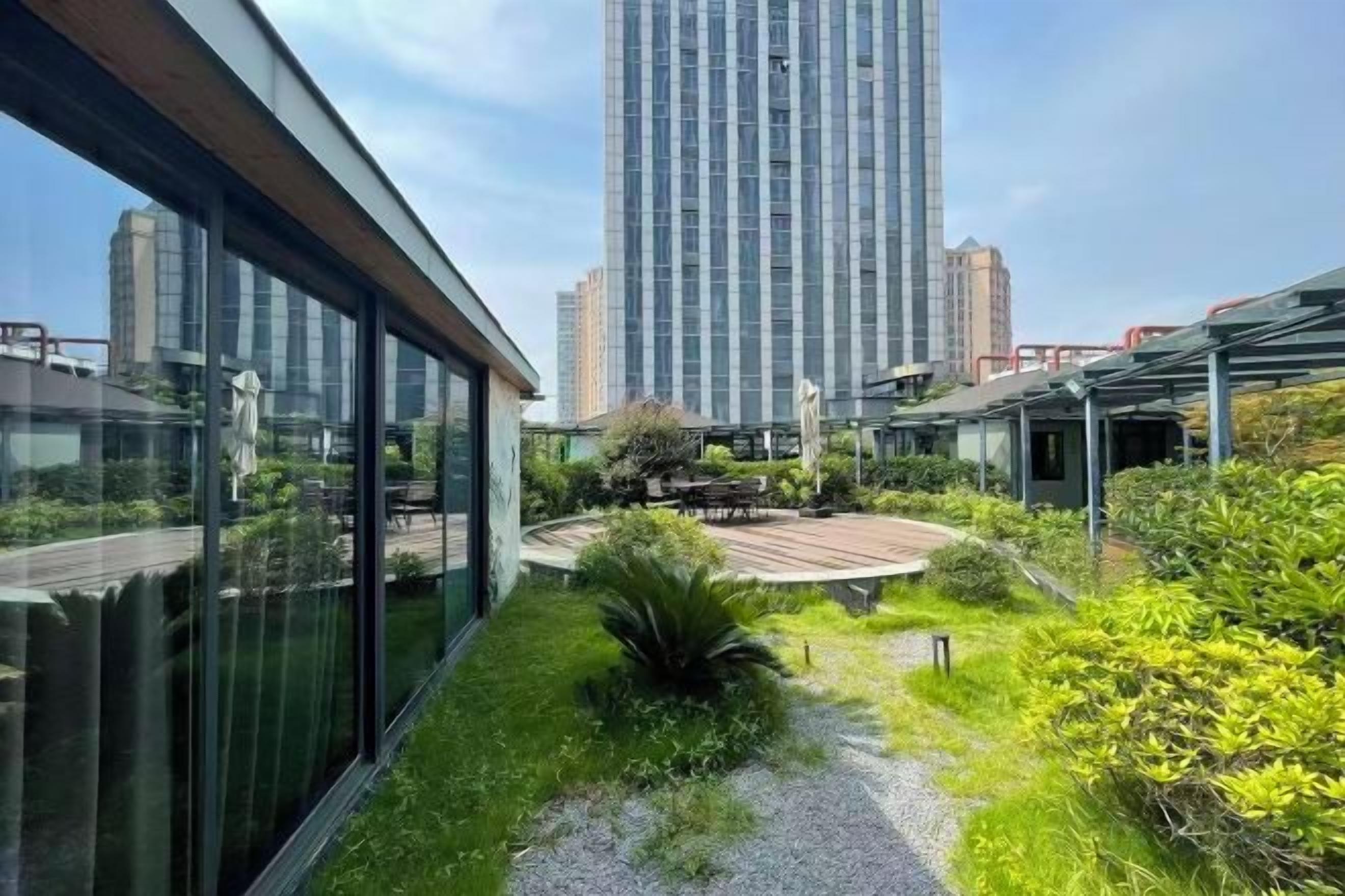 A modern building with a rooftop garden, large windows, and a view of a tall skyscraper.