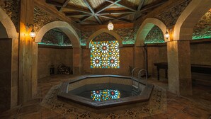 A stone-walled room with a wooden ceiling, a large stained glass window, and a central hot tub.