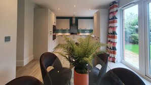A modern kitchen with white cabinets, a central island, and a dining area with two chairs and a potted plant. - Stylish Haven Modern 4-Bedroom Retreat in South Luton with Convenient Parking (Luton)