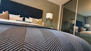 A modern hotel room with a large bed, a headboard, a bedside table with a lamp, and a mirror. - Stylish Haven Modern 4-Bedroom Retreat in South Luton with Convenient Parking (Luton)