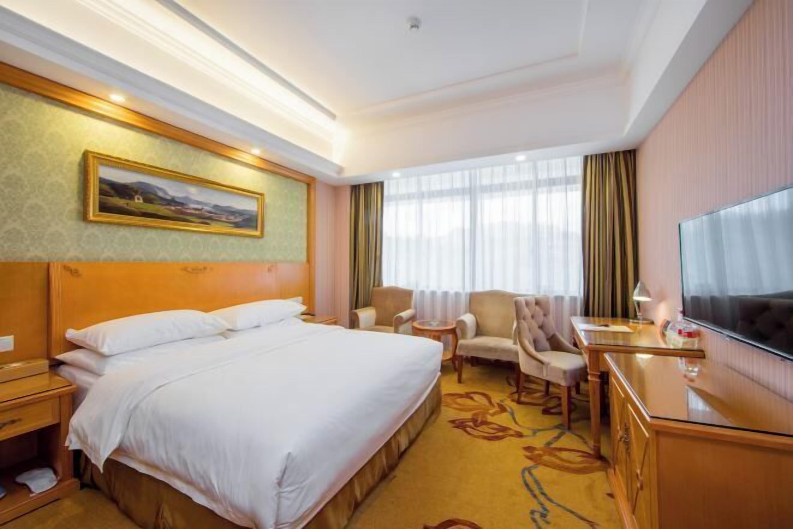 A hotel room with a bed, a desk, a chair, a TV, and a painting on the wall.
